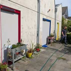 Drei Türen mit bunten Rahmen und Pflanzgefäßen auf einer Terrasse, Mann mit Gartenschlauch.
